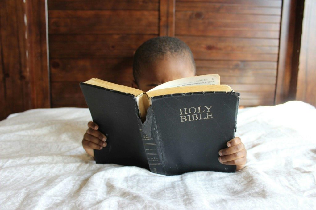 A small child reads from the Bible in bed.