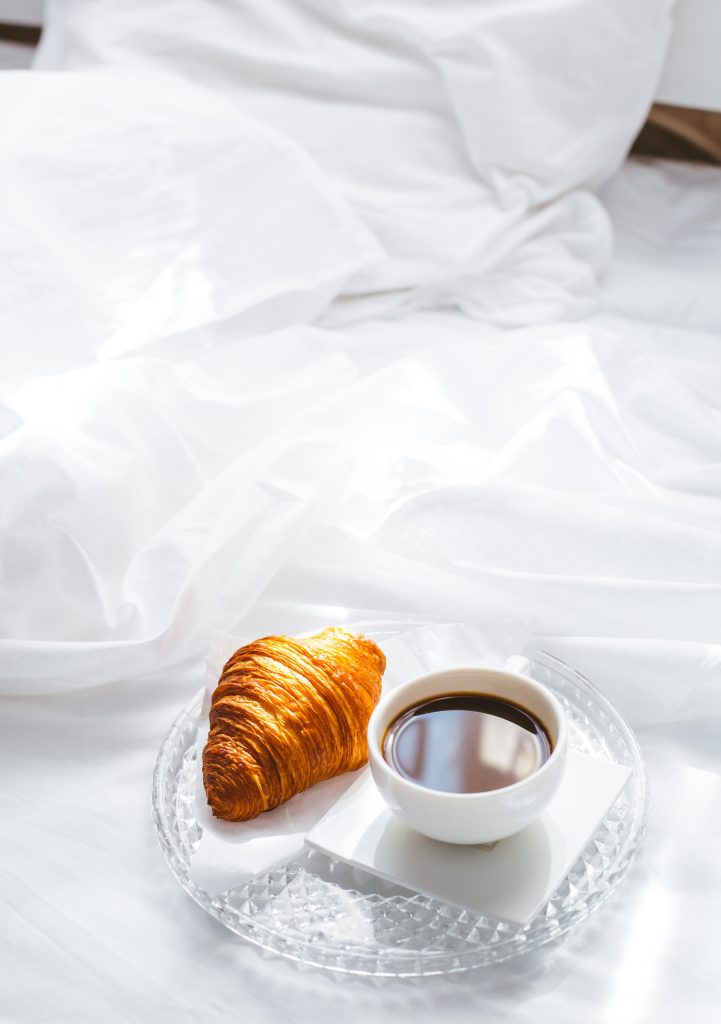 Image of a coffee in a small white cup, next to a golden croissant on a clear glass tray. The tray is on a white surface.
