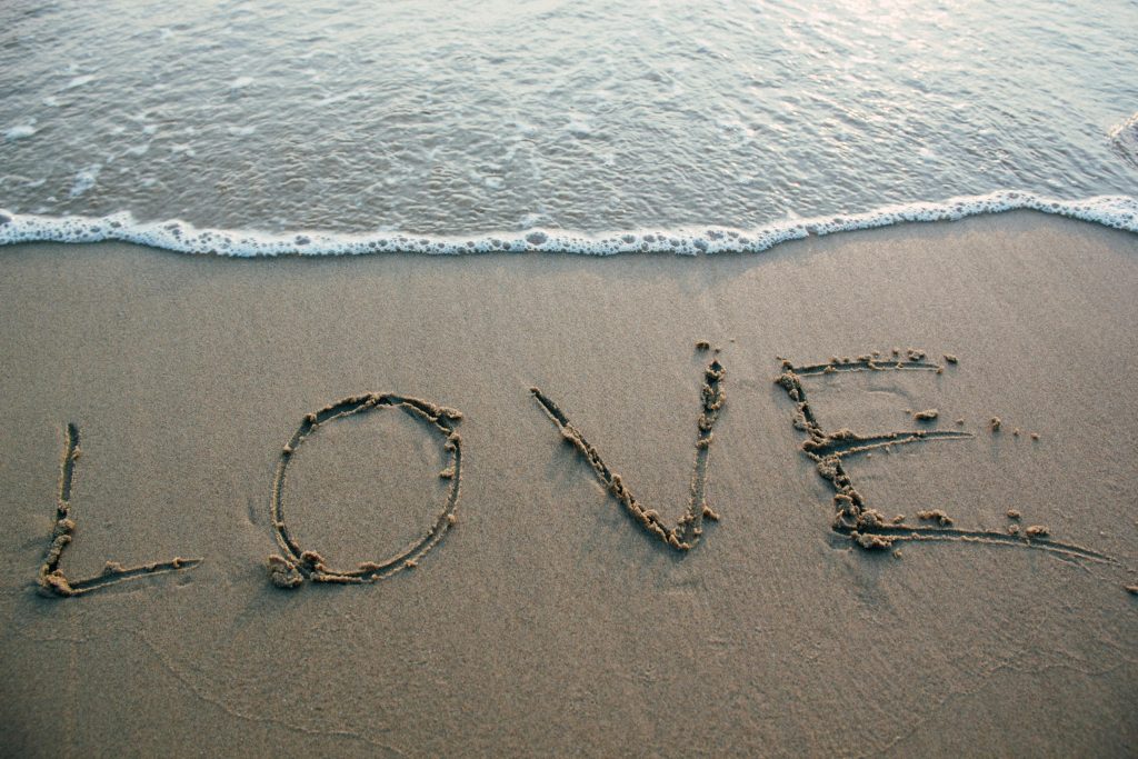 The word "Love" is written in the sand right at the edge of gentle water.