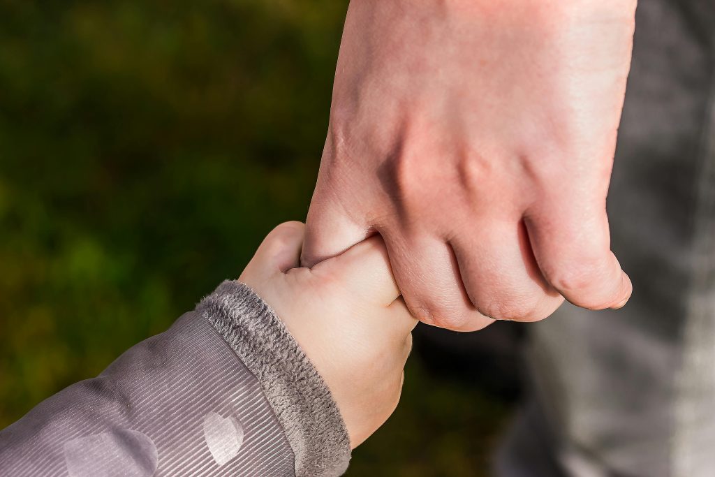 The hand of a toddler is gripping the index finger of an adult in a way that suggests that the child is trusting the adult for guidance.